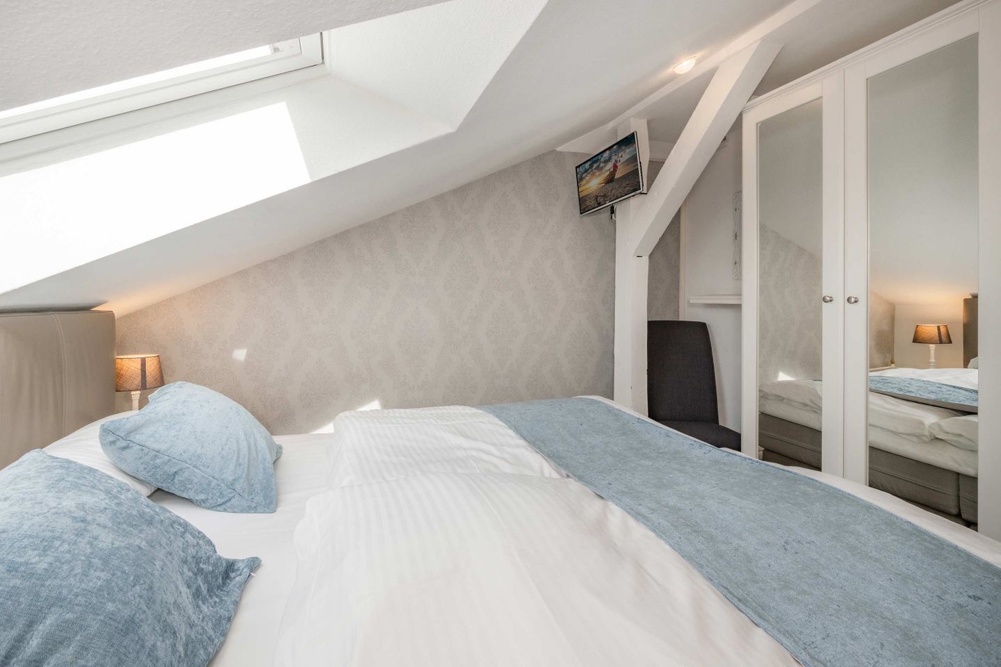 Een slaapkamer met een schuine plafond, een bed met blauw beddengoed, een stoel en een kledingkast.