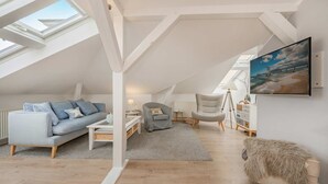 Quarto estilo loft com pé direito alto, janelas no teto, sofá cinza, poltrona cinza, uma mesinha, abajur, um banquinho forrado e um quadro na parede.