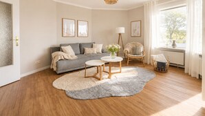 Ein Wohnzimmer mit grauem Sofa, einem runden Couchtisch, einem Korbstuhl und einem großen Fenster mit Vorhängen.