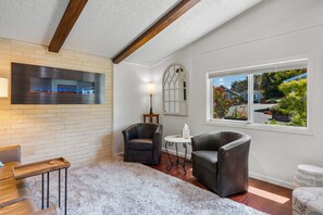 A room with a fireplace, two leather chairs, a small table, and a window with a view of a residential area. - Monarch Pines - Pacific Grove Home - Over 55 Park - Steps to the Sand! (Pacific Grove)