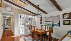 A dining area with a wooden table and chairs, a chandelier, and a view of the outside through a window. - Monarch Pines - Pacific Grove Home - Over 55 Park - Steps to the Sand! (Pacific Grove)