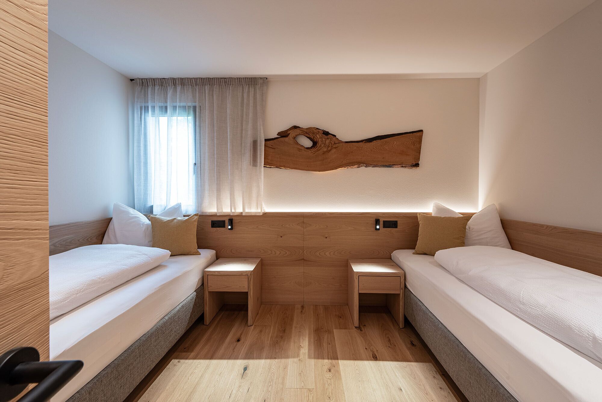 Een moderne hotelkamer met twee enkele bedden, een houten hoofdeinde, een groot raam met dunne gordijnen en een uniek kunstwerk aan de muur.
