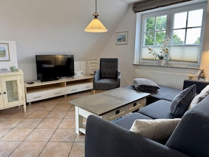 Ein Wohnzimmer mit Fernseher, Sofa, Sessel und einem Fenster mit Blick auf Bäume.
