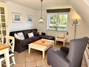 A living room with a sofa, a coffee table, a chair, a cabinet with glass doors, and a window with a view of greenery. - Norderstraße, Apartment 3 - Cozy upper-floor vacation apartment with a south-facing balcony in (Sylt)