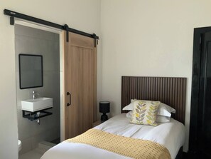 Un dormitorio con cama, una mesita de noche con lámpara, un armario de madera y un baño con lavamanos y espejo.
