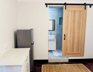 Una cocina con refrigerador, una puerta de madera que da a un baño con lavamanos y espejo.