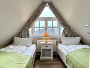 A bedroom with two beds, a nightstand with a lamp, a window with curtains, and a radiator. - Eidumweg Apartment 5 - Bright and modern upper-floor apartment for 4 people with (Sylt)