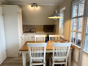 A kitchen with white cabinetry, a wooden dining table, two white chairs, a hanging light fixture, and a window with a brick wall view. - Eidumweg Apartment 5 - Bright and modern upper-floor apartment for 4 people with (Sylt)