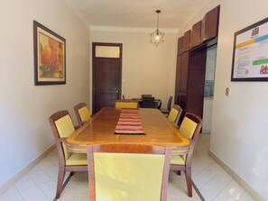 Comedor con mesa de madera, sillas amarillas, alfombra, pintura, puerta y cocina visible a través de una abertura.