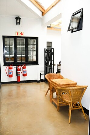 A room with a large window, two fire extinguishers, a wicker table, and a chair. - Double Self-Contained Rooms 2 (Johannesburg)