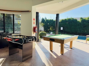 A pool table and foosball table in a well-lit room with large windows.