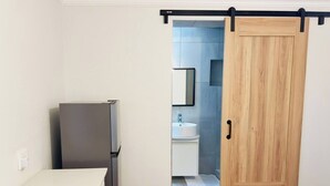 Una cocina con refrigerador, una puerta de madera que da a un baño con lavamanos y espejo.