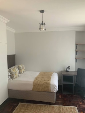 A single bed with a headboard, a bedside table with a lamp, a chair, and a woven rug on a wooden floor.