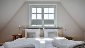 Et soveværelse med skrå væg, et stort vindue med persienner, to sengeborde med lamper og en seng med et gråt tæppe.