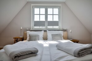 Ein Schlafzimmer mit schrägen Wänden, einem großen Fenster mit Jalousien, zwei Nachttischen mit Lampen und einem Bett mit grauer Decke.