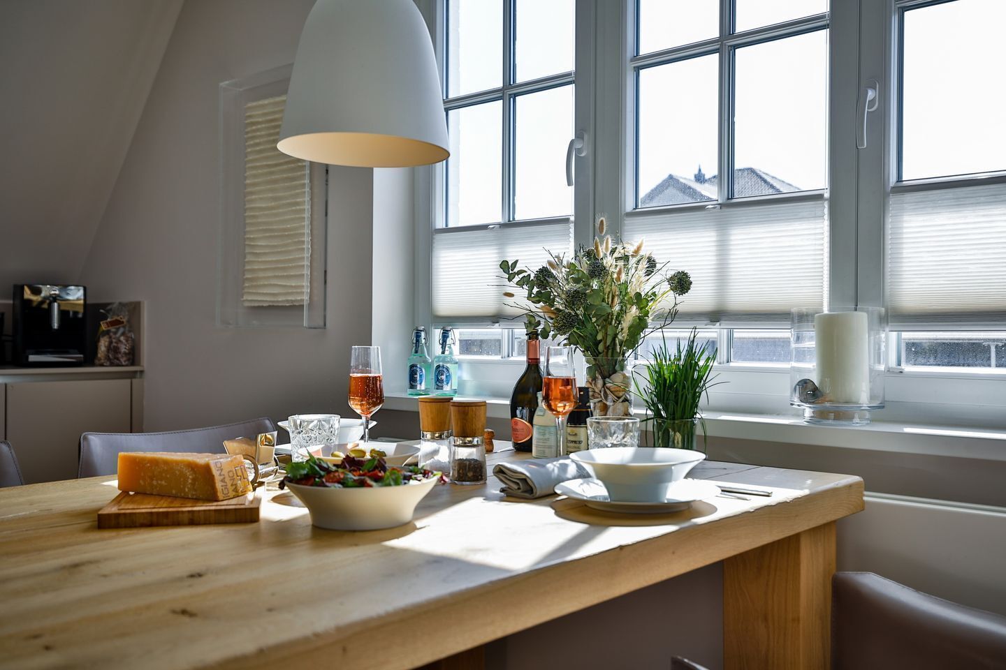 A dining area with a wooden table, various food items, glasses, and a vase with flowers.