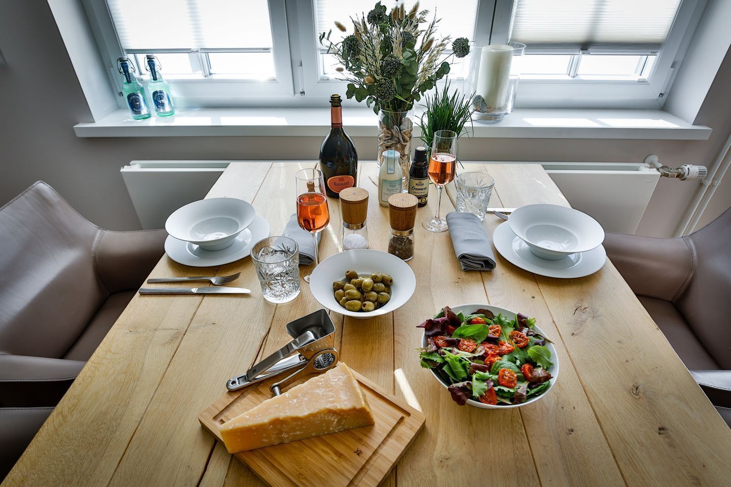 Une table à manger dressée avec des assiettes, des verres, de l’huile d’olive, une bouteille, un verre de vin, un saladier et une planche à fromages.