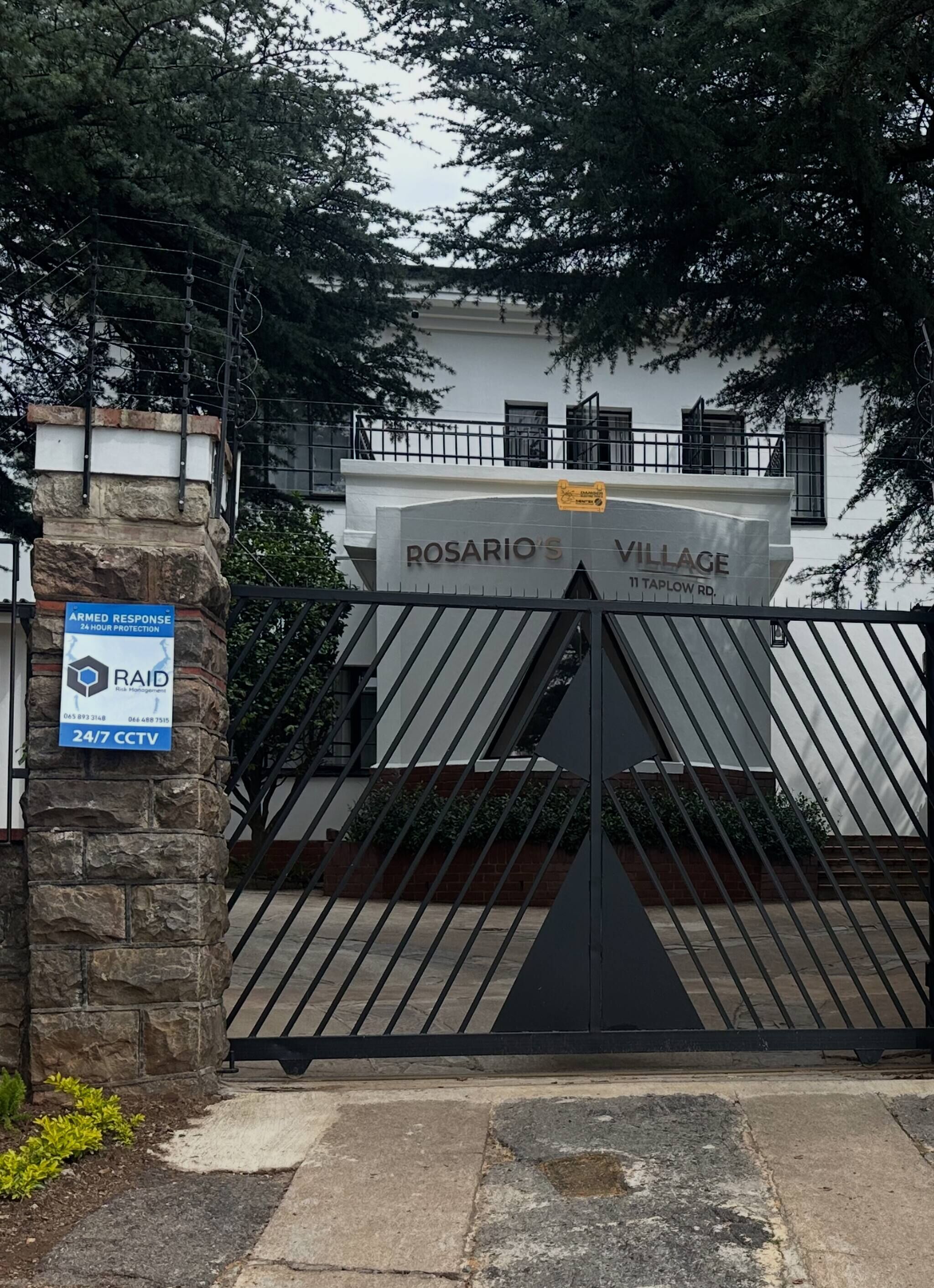 Une entrée fermée par un portail mène à un immeuble appelé « Rosario’s Village », avec un pilier en pierre et une pancarte indiquant la présence d’une équipe d’intervention armée et d’un système de vidéosurveillance fonctionnant 24 heures sur 24, 7 jours sur 7.