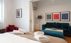 Ein Hotelzimmer mit einem Bett, einem roten Sessel, einem türkisgrünen Sofa und drei gerahmten Kunstwerken an der Wand.