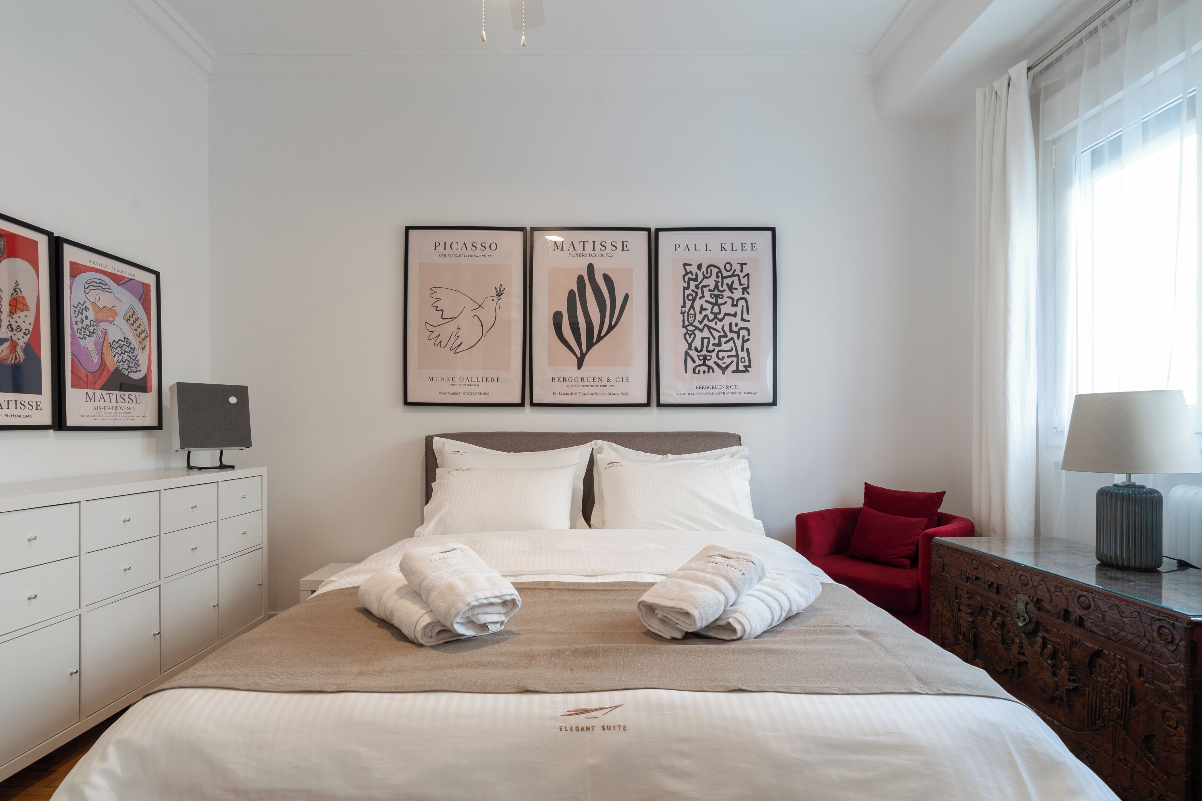 A hotel room with a neatly made bed, a red chair, and three framed artworks on the wall.