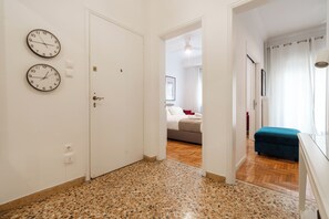 A hallway with a white door, two wall clocks, and a glimpse of a bedroom with a bed and a blue chair. - Elegant Pangrati Apartment Near Metro & Museums 
Entire apartment Up to 4 people (Athina)