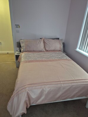 A bedroom with a bed, two bedside tables, a nightstand, a wall-mounted light switch, and a window with blinds.