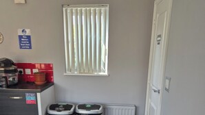 A compact living space with a kitchenette, a grey sofa, two trash bins, and a window with blinds. - Charming Guest Room in West Midlands with Modern Amenities (West Midlands)