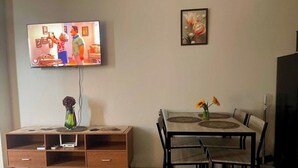 A living room with a TV mounted on the wall, a dining table with chairs, a wooden cabinet with drawers, and a vase with flowers on the table. - Cozy Retreat by Jane (Cebu City)