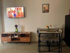 Ein Wohnzimmer mit Wandfernseher, einem Esstisch mit Stühlen, einer Holzkommode mit Schubladen und einer Vase mit Blumen auf dem Tisch.