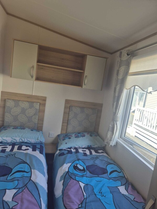 A bedroom with two beds, each with a cartoon character print, a wooden shelf above, and a window with curtains.