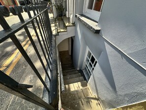 Eine Treppe führt zu einer Empfangsdiele im zweiten Stock, mit einem Metallgeländer, einem Topf mit Pflanze und einem Fenster mit weißem Rahmen.