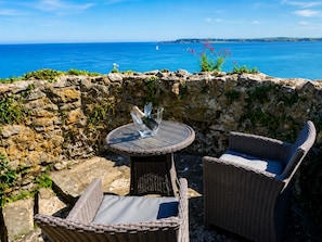 Ein Sitzbereich im Freien mit Korbmöbeln, einem Tisch und Stühlen, mit Blick auf die Küste, das Meer und das entfernte Land.