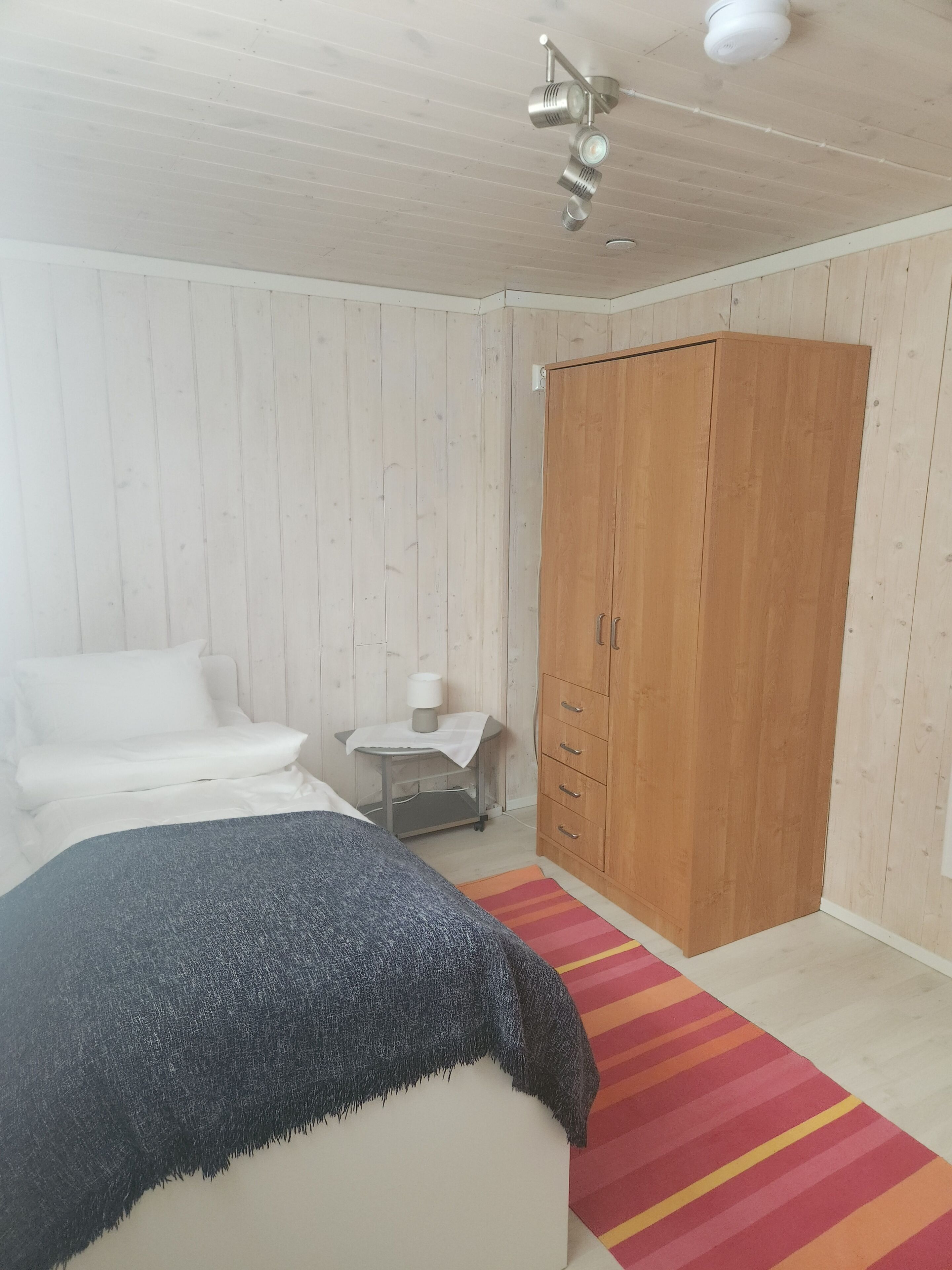 Ein Schlafzimmer mit einem Bett, einem Kleiderschrank, einem Nachttisch mit Lampe und einem Streifen Teppich.