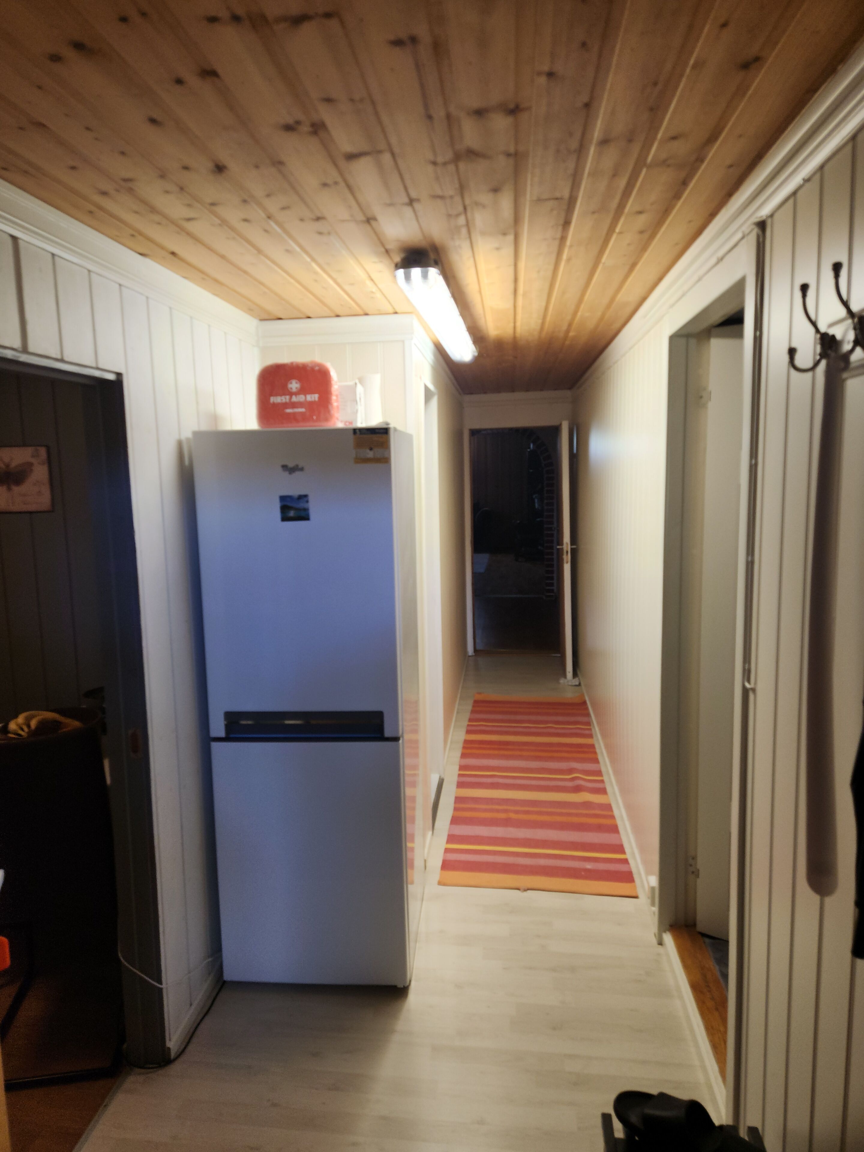 Een smalle gang met een houten plafond, een witte koelkast, een gestreept tapijt en een deur met kapstok.