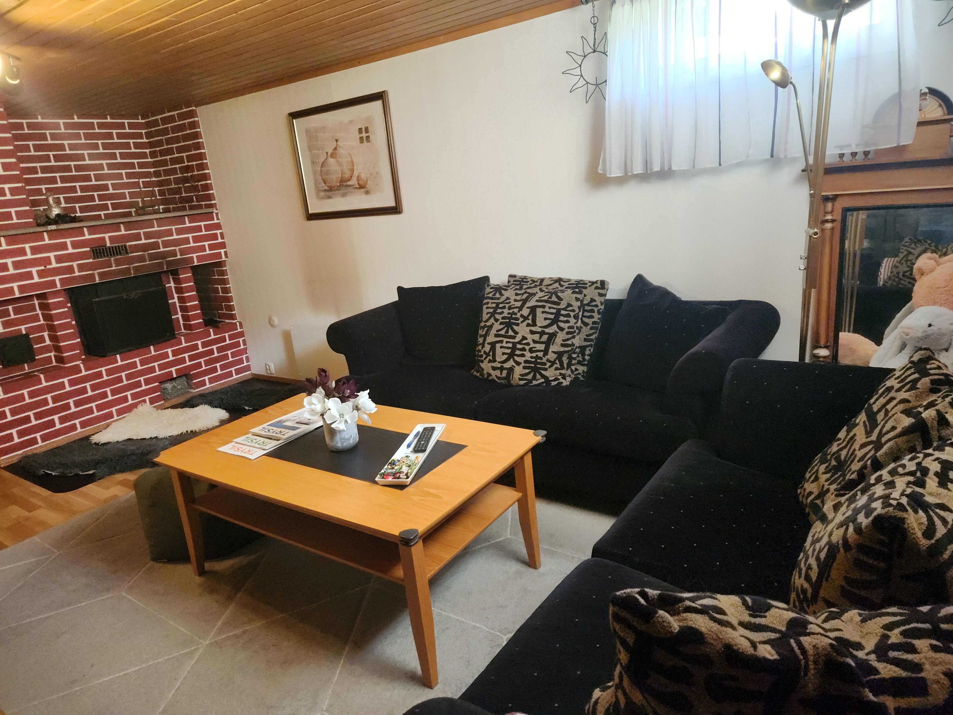 Een woonkamer met een open haard van baksteen, een zwarte bank met gekleurde kussens, een houten salontafel en een kunstwerk in een lijst aan de muur.