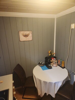 A small room with a round table covered with a white tablecloth, a chair, a wall-mounted picture, and various items on the table including a vase with flowers, a coffee maker, and some bottles.