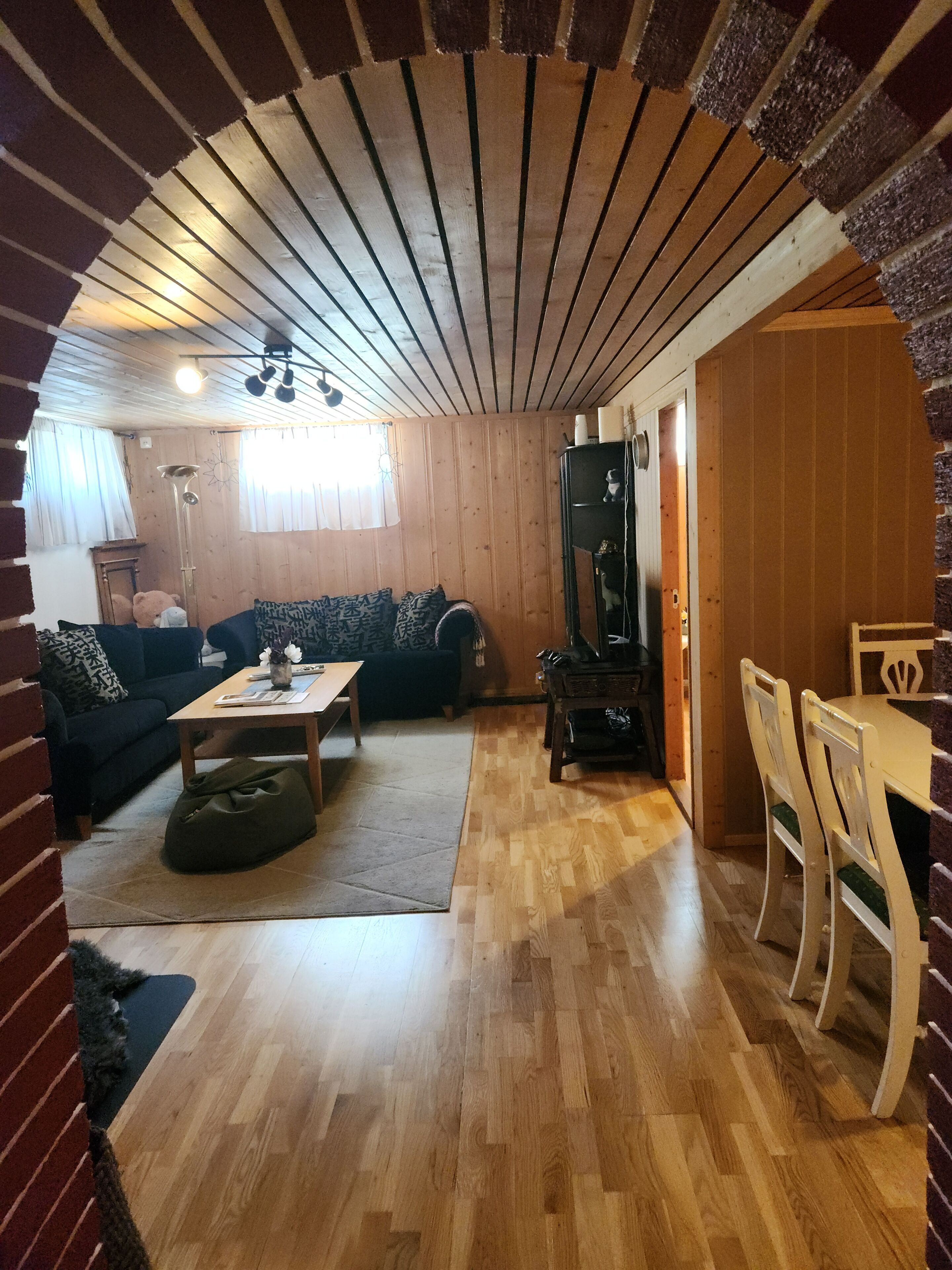Ett mysigt vardagsrum med ett träpanelat tak, en soffa, ett soffbord och en matplats med stolar.