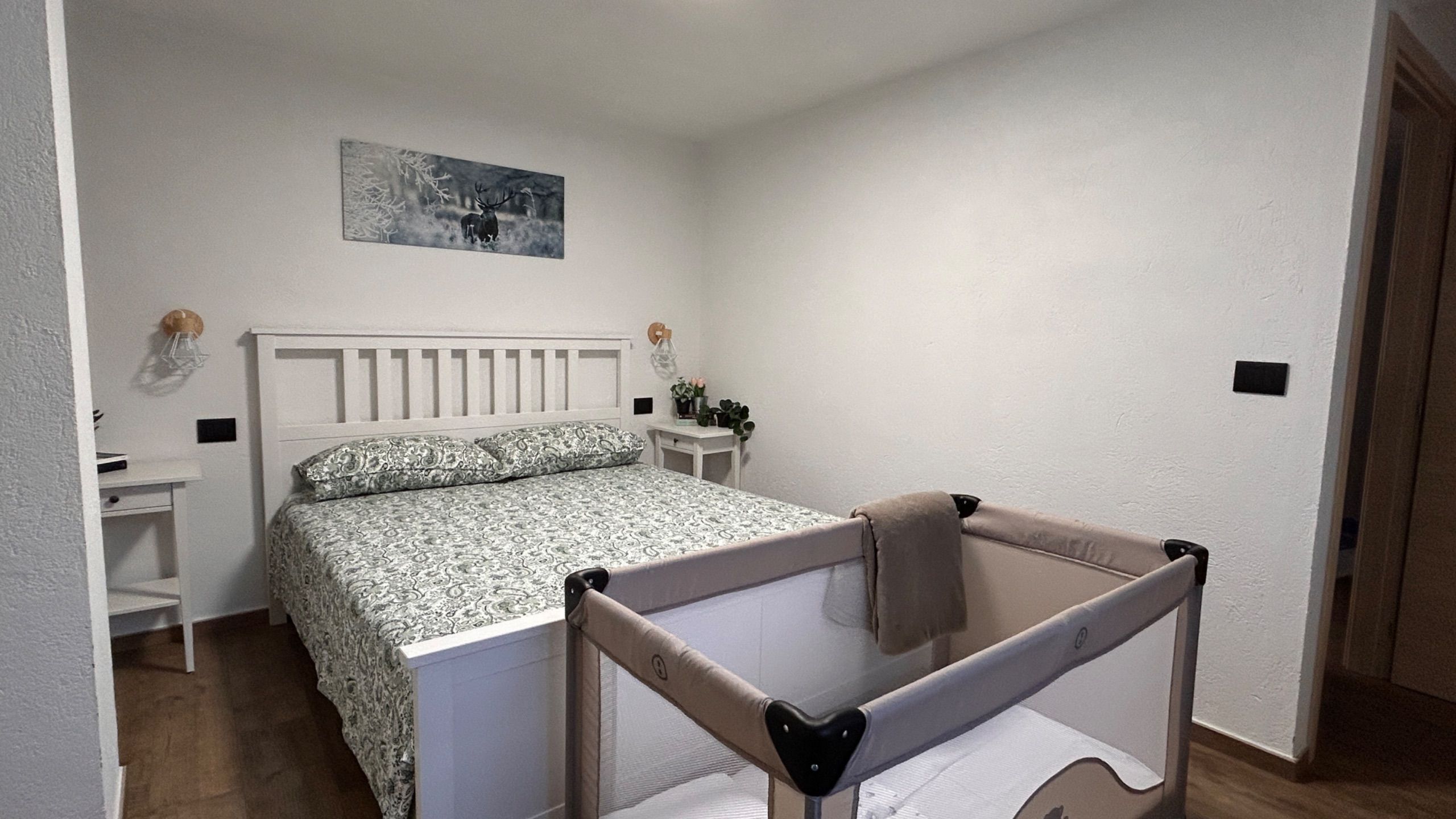 Een slaapkamer met een bed, nachtkastje, bijzettafeltje, ledikant en een aan de muur hangend schilderij.