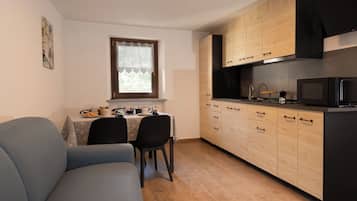 Ein kompakter Wohnraum mit einer Couch, Esstisch, Stühlen, einer Küchenzeile mit Schränken und Geräten sowie einem Fenster mit Vorhang.