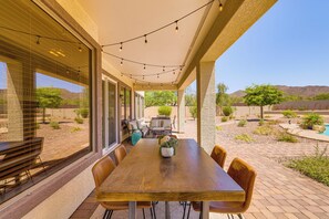 Un patio exterior con mesa y sillas de madera, luces colgantes y vistas a una piscina y paisaje desértico.
