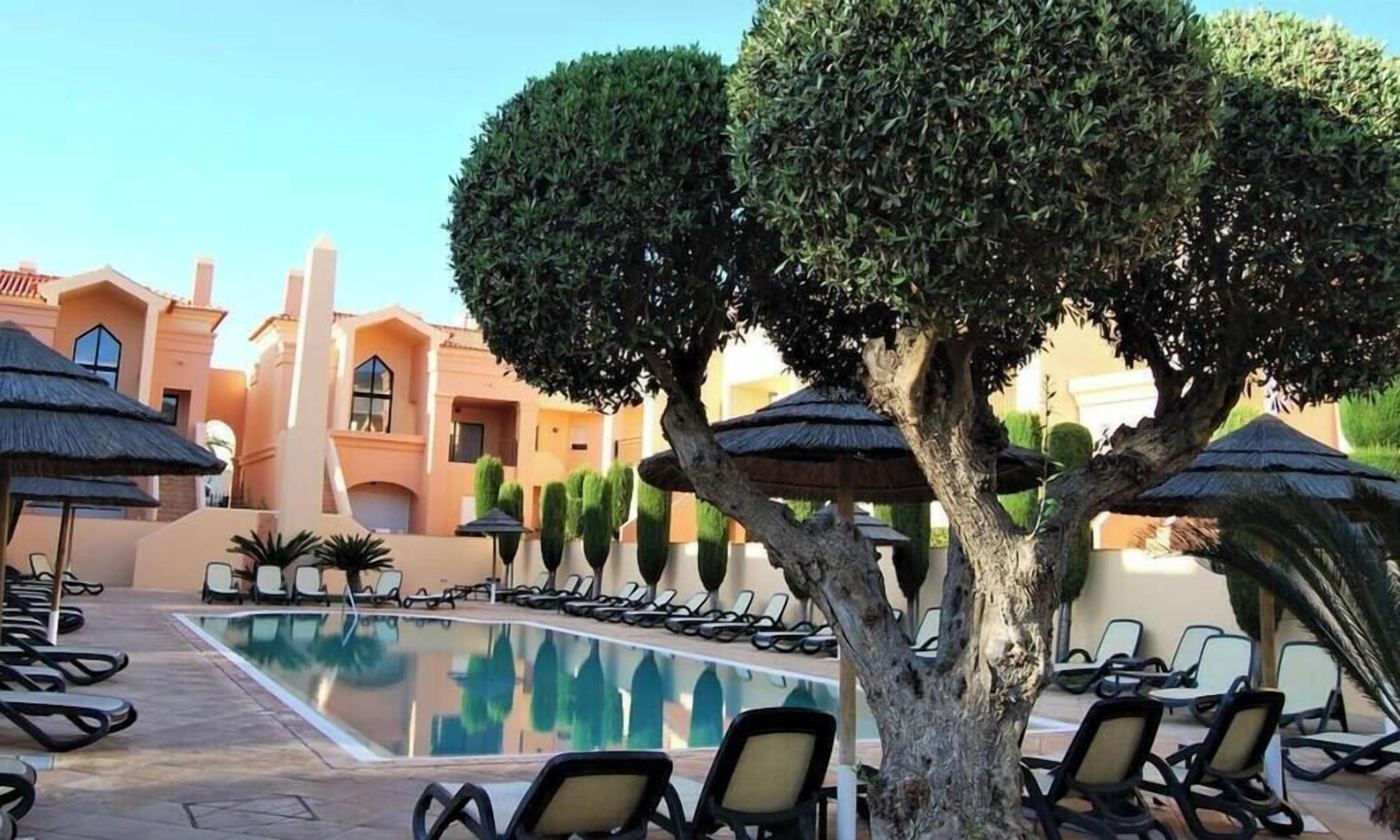 A poolside area with lounge chairs and umbrellas, surrounded by neatly trimmed hedges and a building with a Mediterranean architectural style.