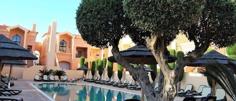 A poolside area with lounge chairs and umbrellas, surrounded by neatly trimmed hedges and a building with a Mediterranean architectural style.