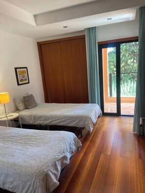 A hotel room with two beds, a wooden wardrobe, a framed picture on the wall, and a balcony with a view of greenery. - Luxurious 2-Bedroom Apartment in Luz, Portugal (Luz)