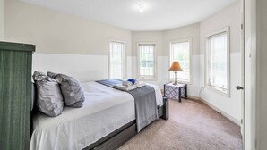 Un dormitorio con cama, almohadas, una mesita de noche con lámpara y ventanas.