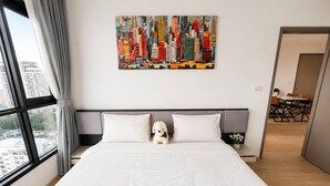 A hotel room with a large bed, a cityscape painting, a window with curtains, and a view of buildings. - Sea View 2 Bed Room Excellent Location (Muang Pattaya)