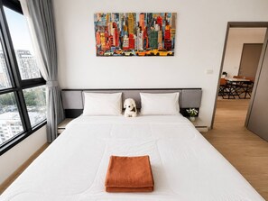 A hotel room with a large bed, a cityscape painting, a window with curtains, and a view of buildings. - Sea View 2 Bed Room Excellent Location (Muang Pattaya)