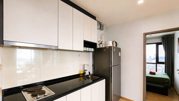 A modern kitchen with white cabinets, a black countertop, a stainless steel refrigerator, and a window with a city view. - Sea View 2 Bed Room Excellent Location (Muang Pattaya)