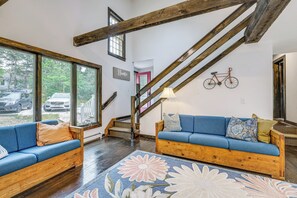 Un salón amplio con una escalera de madera, un conjunto de sofá azul y un ventanal con vistas a coches aparcados y árboles.