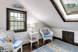 Una habitación con dos sillones de mimbre, una mesita con lámpara, una ventana y una alfombra estampada.