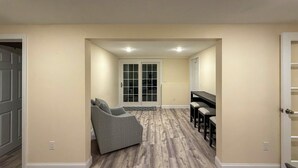 A hallway with a gray sofa, a dining area with a table and chairs, and a door leading to a room. - Minutes Away From the Best Beaches in Cape Cod! (South Dennis)